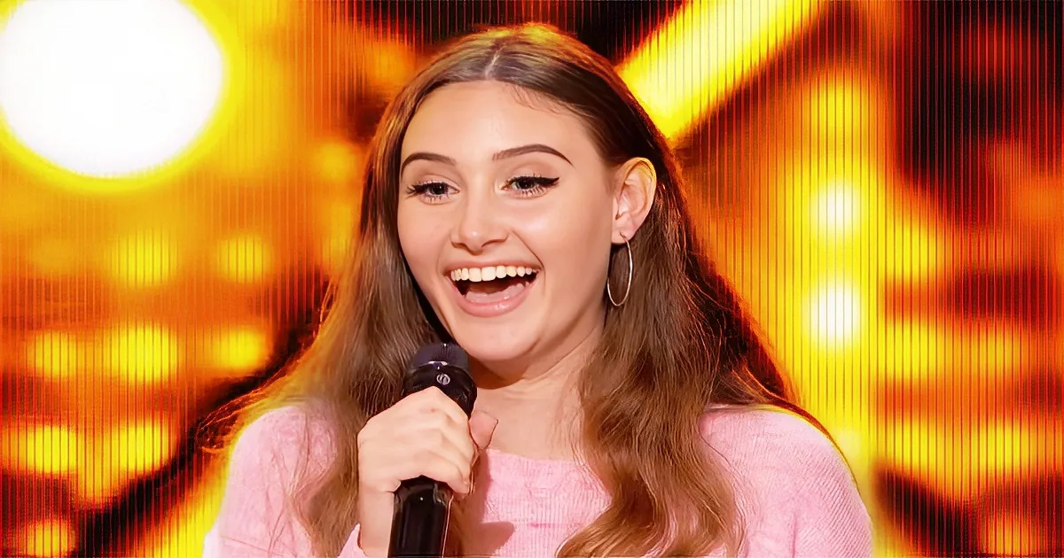 A person with long hair, wearing a pink top, smiles while holding a microphone against a vibrant, golden background.