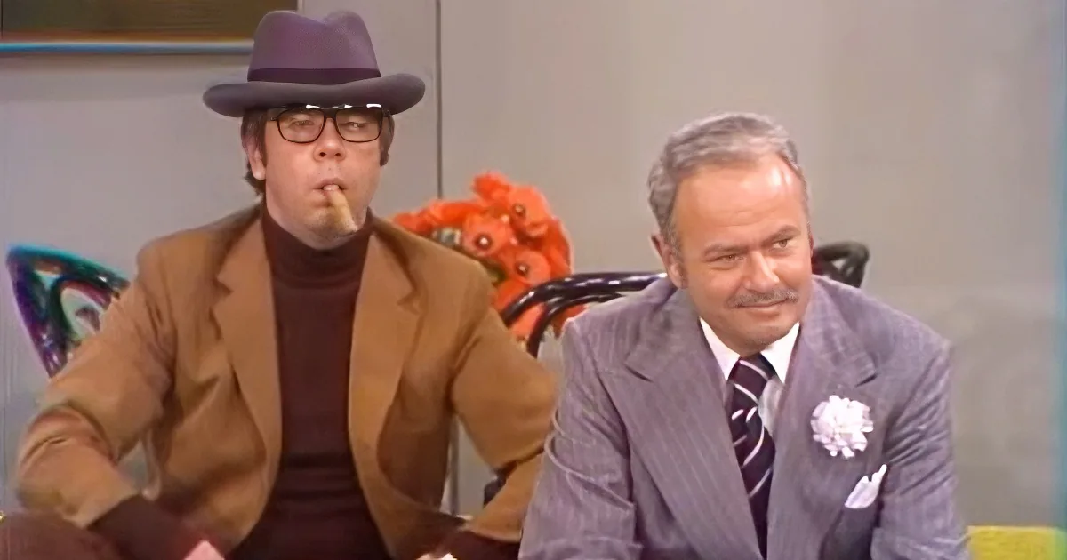 A man in a brown suit and hat smokes a cigar, sitting next to another man in a gray suit with a white flower on his lapel.