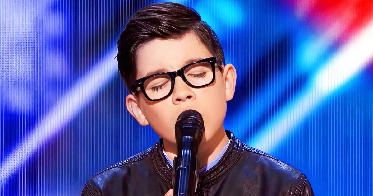 A young boy wearing glasses and a black leather jacket sings into a microphone on a stage with blue lighting.