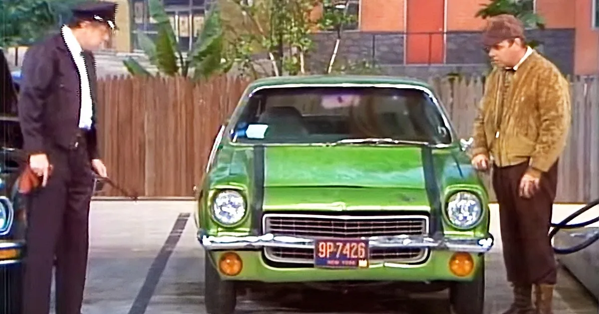 A green car with a New York license plate is parked, with two men standing nearby, one in a uniform and the other in a...