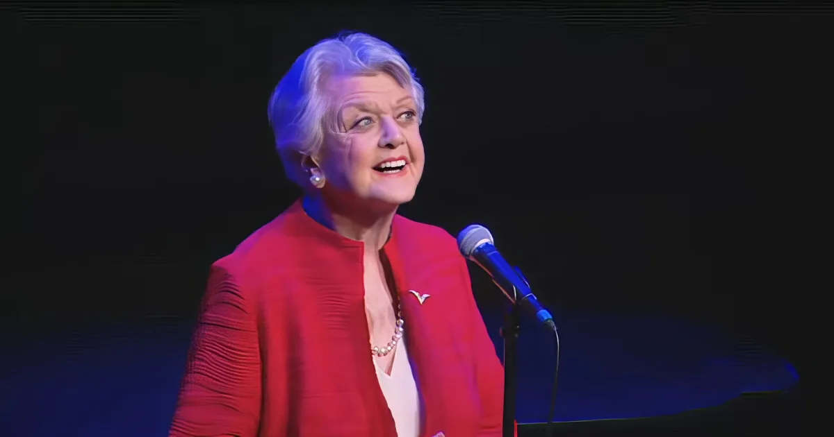 An older woman in a red outfit speaks into a microphone on stage.