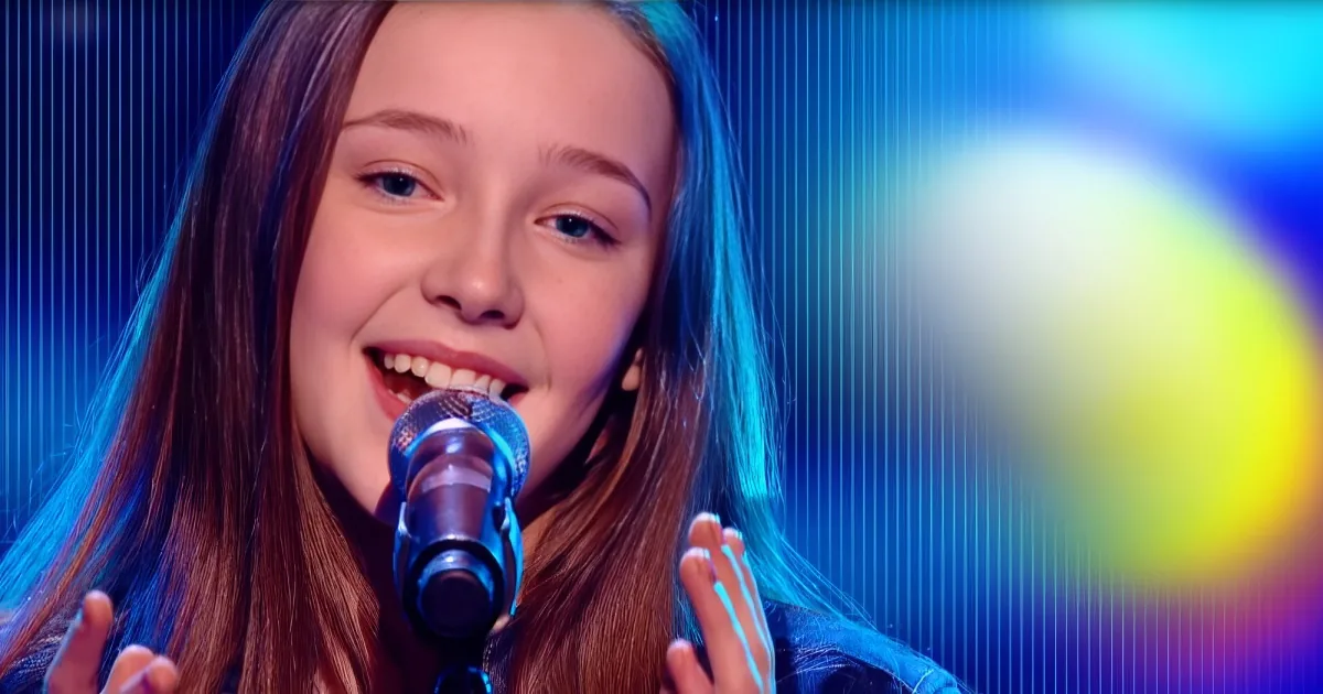 A young girl with long brown hair sings into a microphone on stage with colorful lighting.