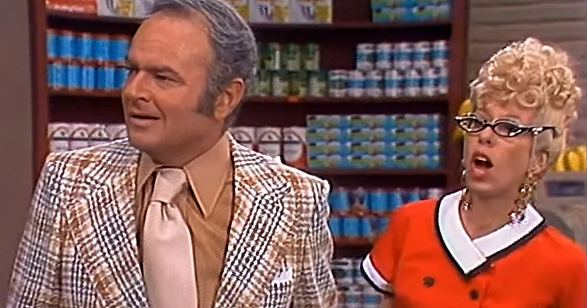 A man in a checkered suit and a woman in an orange dress with glasses are in a store setting.
