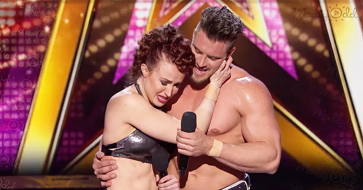 A man and woman are embracing on stage, both shirtless, with the woman holding a microphone.