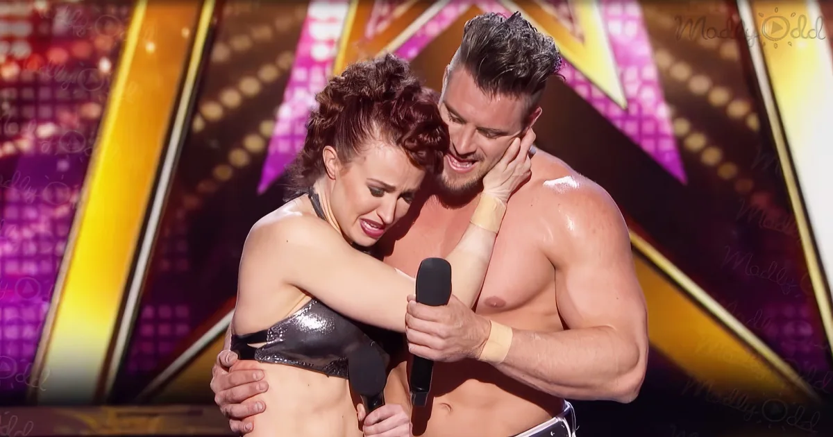 A man and woman are embracing on stage, both shirtless, with a microphone in the woman's hand.