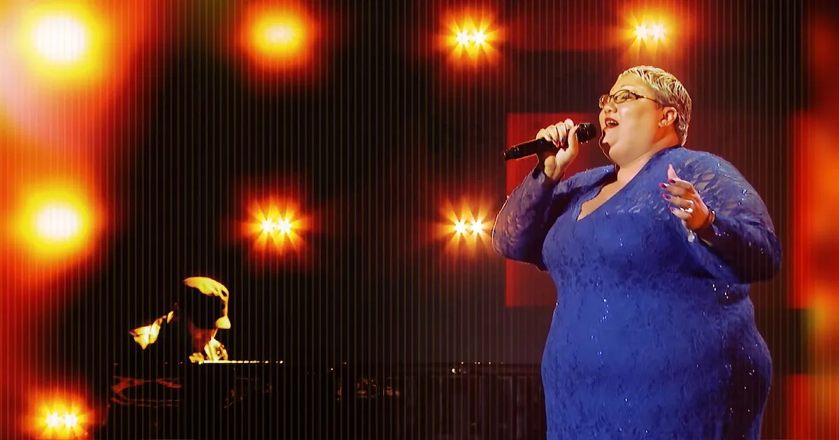 A person in a blue dress sings into a microphone on stage with bright lights.