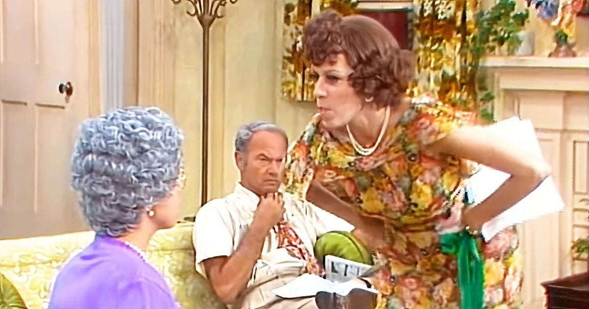 A woman in a floral dress stands with hands on hips, talking to another woman in a purple top, while a man sits on a...