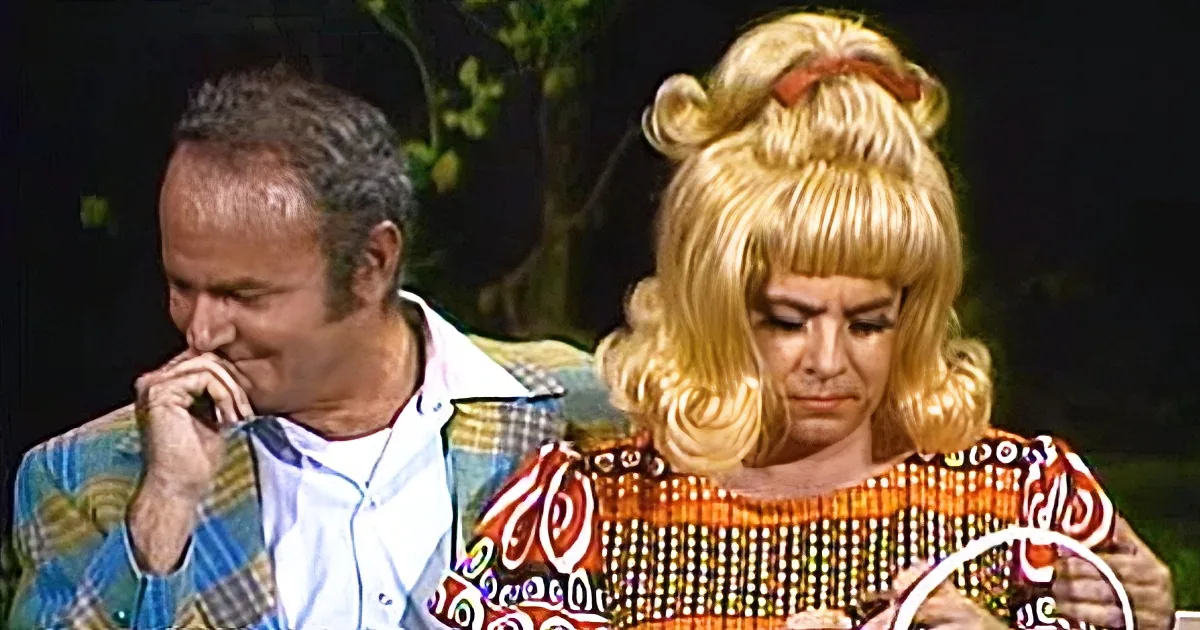 A man in a plaid jacket and a woman in a colorful dress are seated, both appearing to be in a conversation or interview...