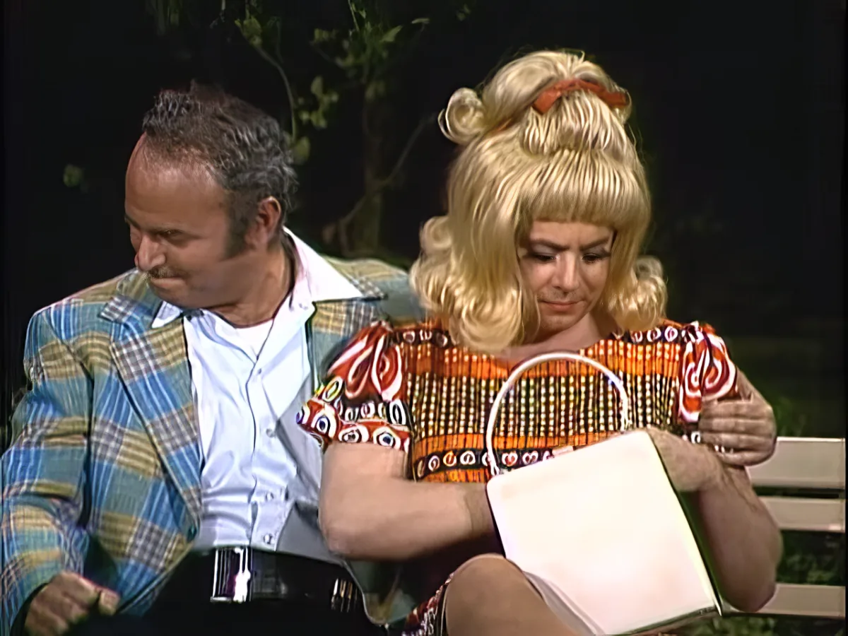 A person in a colorful outfit holds a stack of orange and white items while another person in a plaid jacket looks on.