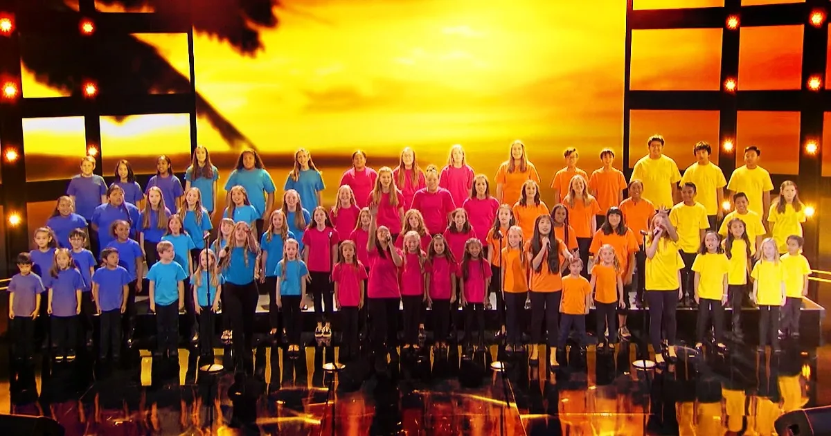 A large choir of children in blue, pink, orange, and yellow shirts stands on stage against a sunset backdrop.