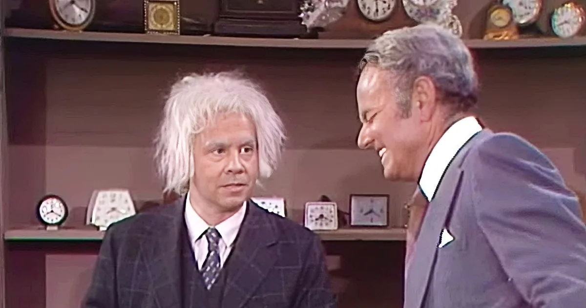 A man in a suit with white hair and another man in a suit laughing, surrounded by clocks on shelves.