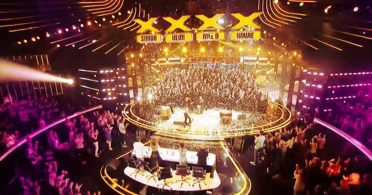 A performer stands on a stage surrounded by a large, enthusiastic crowd under vibrant stage lighting.