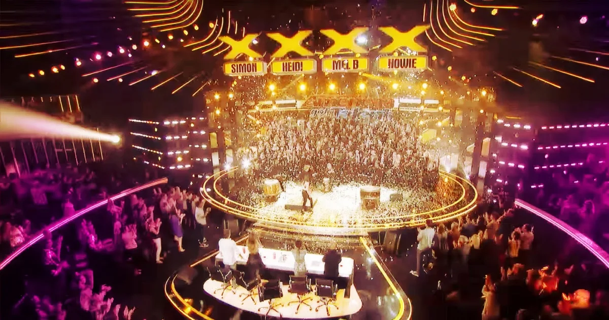 A vibrant stage with judges' names displayed, surrounded by a cheering crowd and bright stage lights.