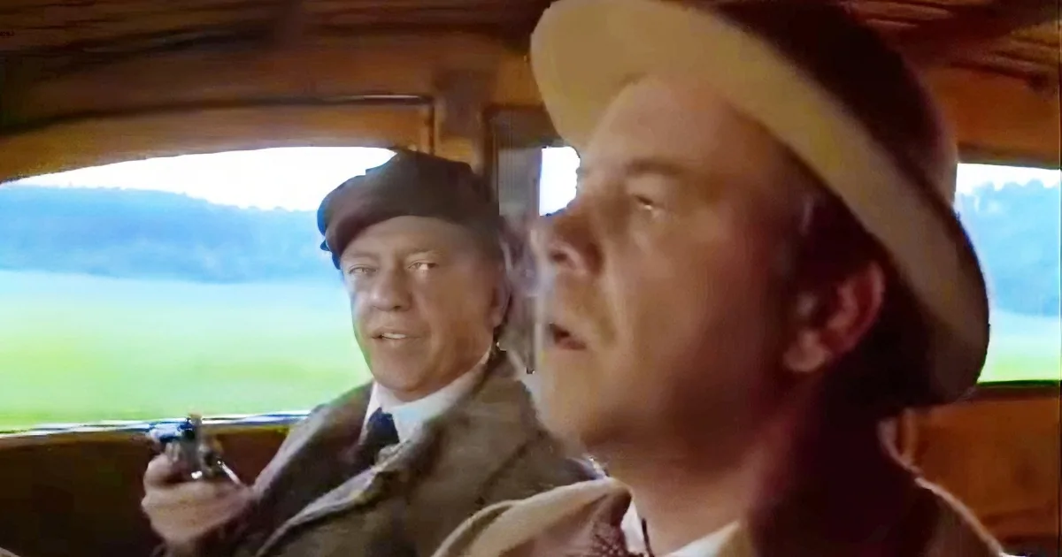 A man in a cowboy hat is driving a car, while another man in a suit sits behind him, holding a gun.
