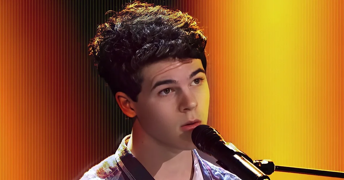 A young man with curly hair sings into a microphone against a vibrant orange background.