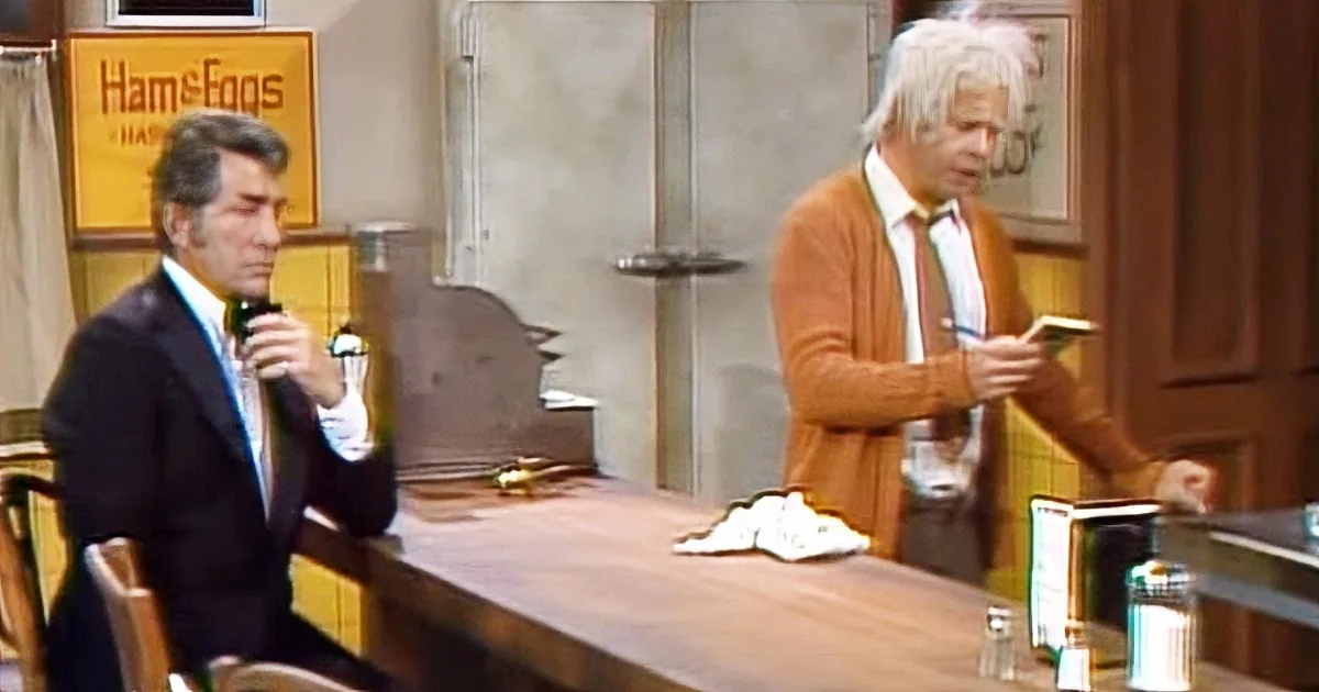 A man in a suit sits at a bar, holding a drink, while another man in a sweater stands nearby, holding a book.