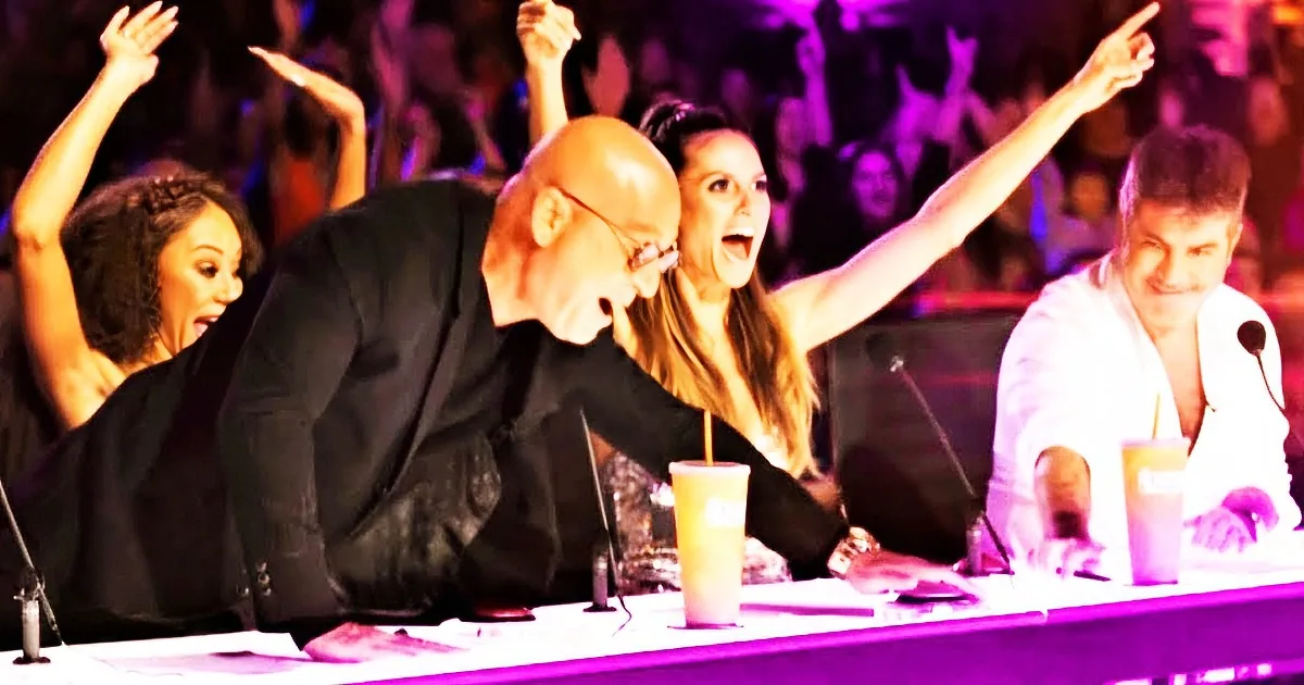 A man leans forward excitedly, surrounded by people raising their arms in celebration at a judging panel.