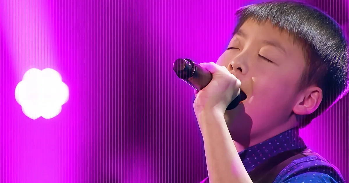 A young boy sings passionately into a microphone under vibrant purple stage lighting.