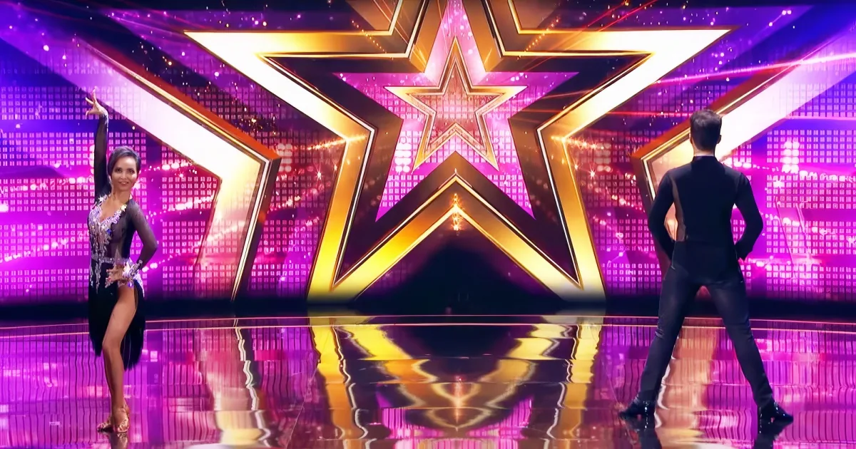 A woman in a black dress stands on a stage with a large golden star backdrop.