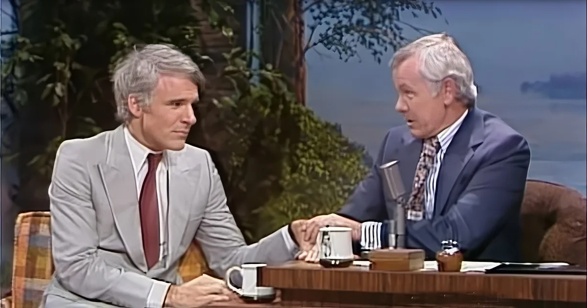 Two men in suits are seated at a table, engaged in conversation on a talk show set.