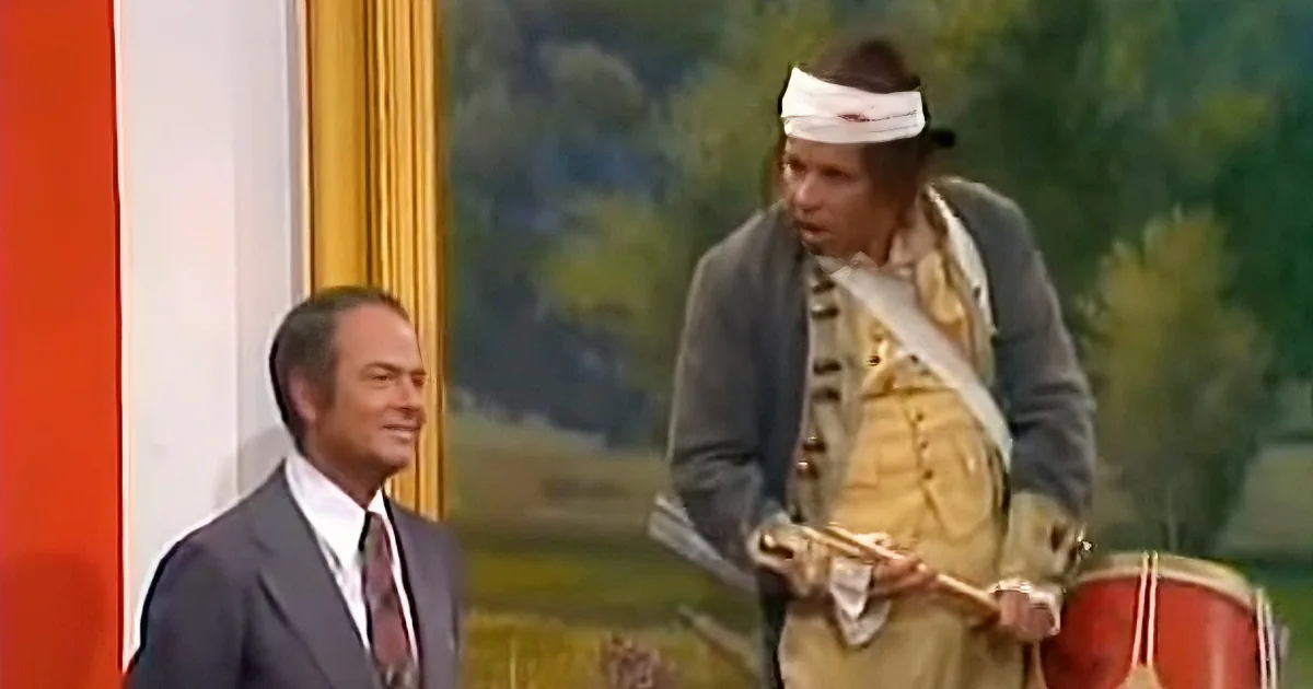 A man in a suit smiles as another in Native American attire plays a drum.