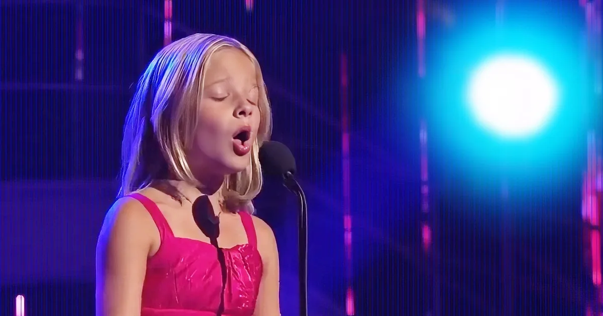 A young girl in a pink dress sings into a microphone on a stage with blue and purple lighting.