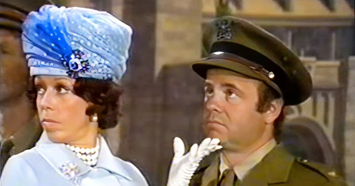 A woman in a blue dress and turban stands beside a man in a military uniform, both looking off to the side.