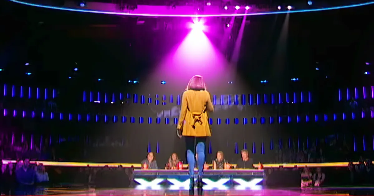 A person in a yellow coat stands on a stage under a bright purple spotlight, facing away from the camera.