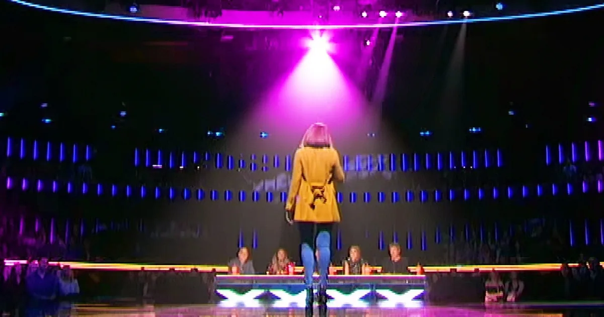 A person in a yellow coat walks onto a brightly lit stage with a spotlight.