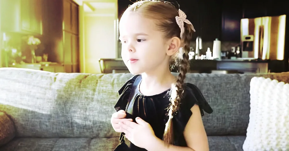 A young girl with braided hair and a bow, wearing a black dress, sits on a couch in a sunlit living room.