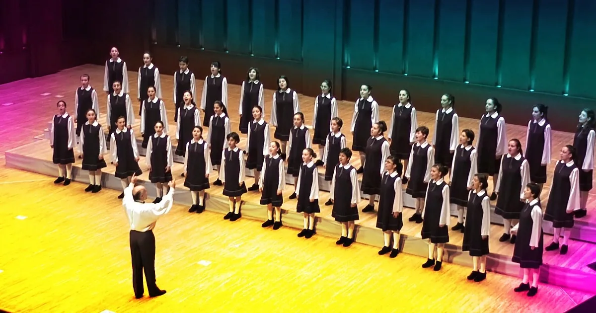 A conductor leads a choir of young girls in uniform on a stage.
