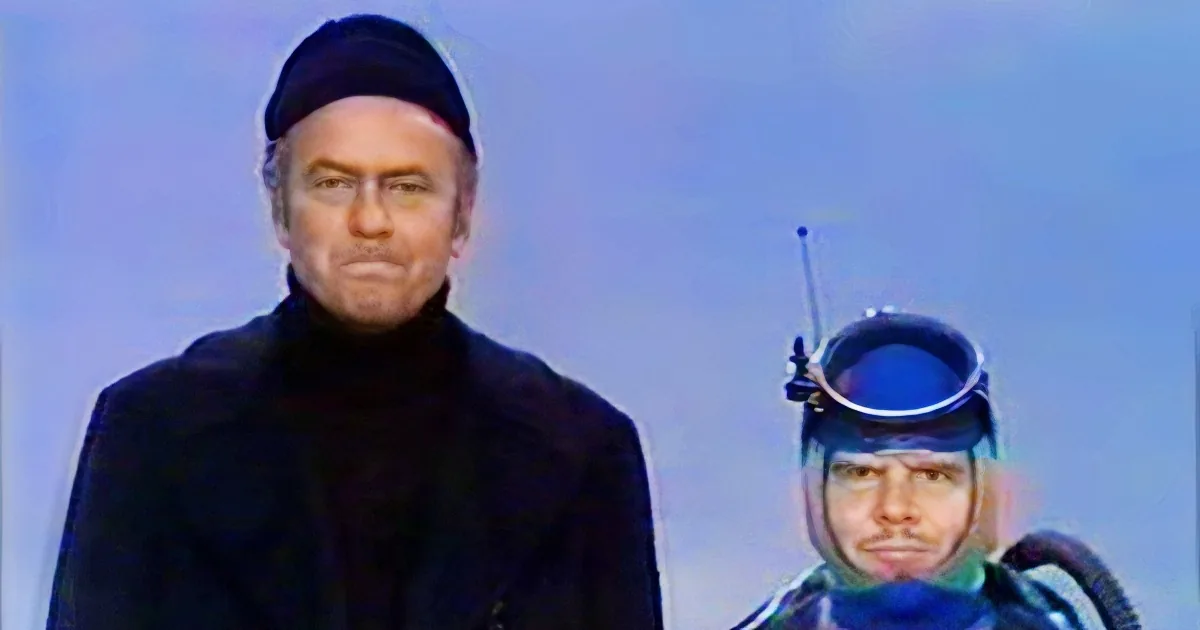 A man in a black outfit and hat stands next to another person wearing a diving helmet and blue gear.