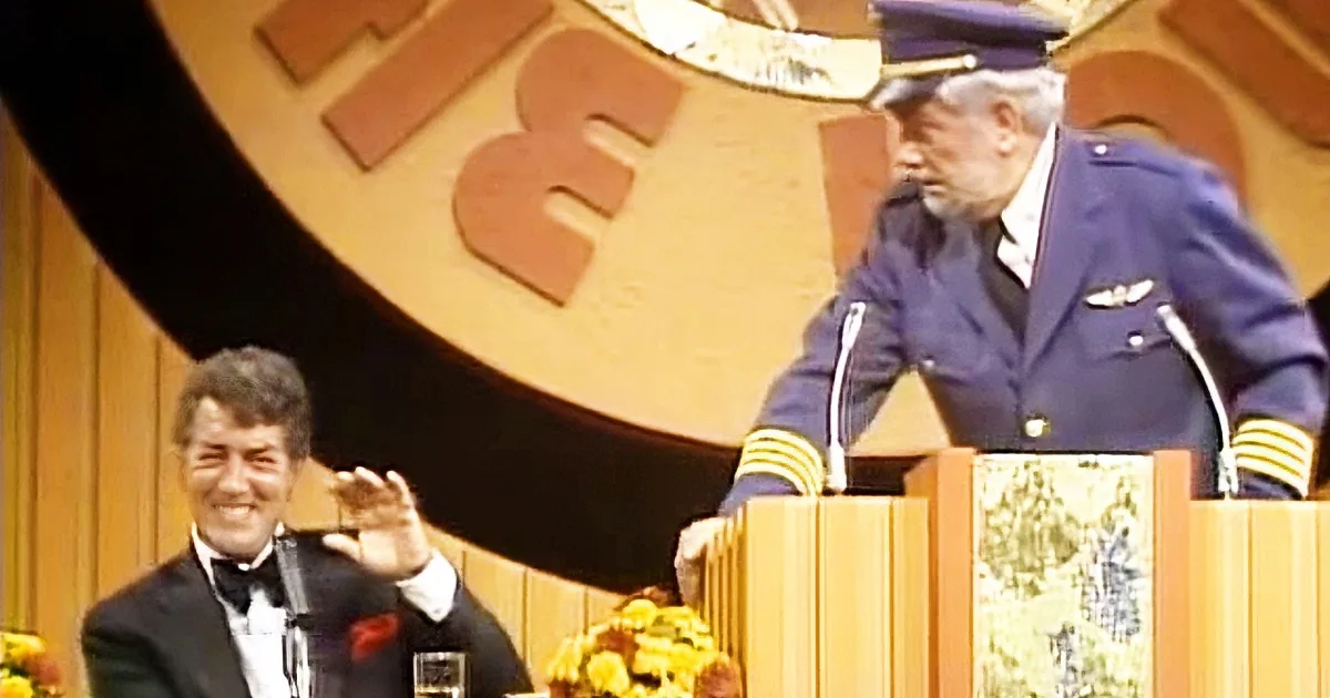 A man in a blue uniform stands at a podium, addressing an audience, while another man in a tuxedo sits and gestures with...