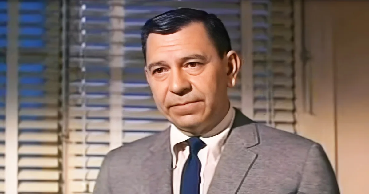A man in a suit and tie stands in front of a window with blinds, looking serious.