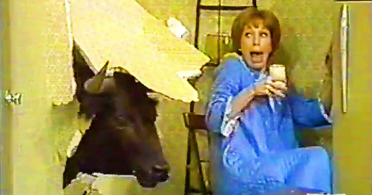 A woman in a blue dress sits on a chair, holding a drink, while a cow's head peeks through a hole in the wall behind her.