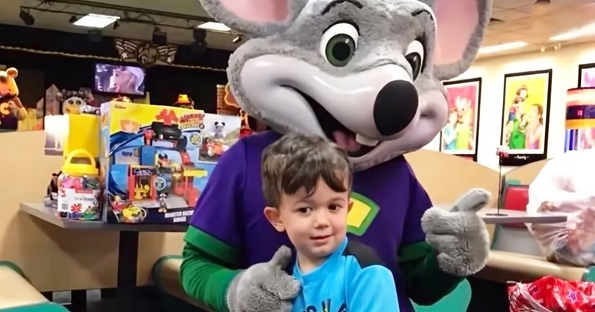 A child sits next to a large gray mouse mascot in a colorful, toy-filled store.