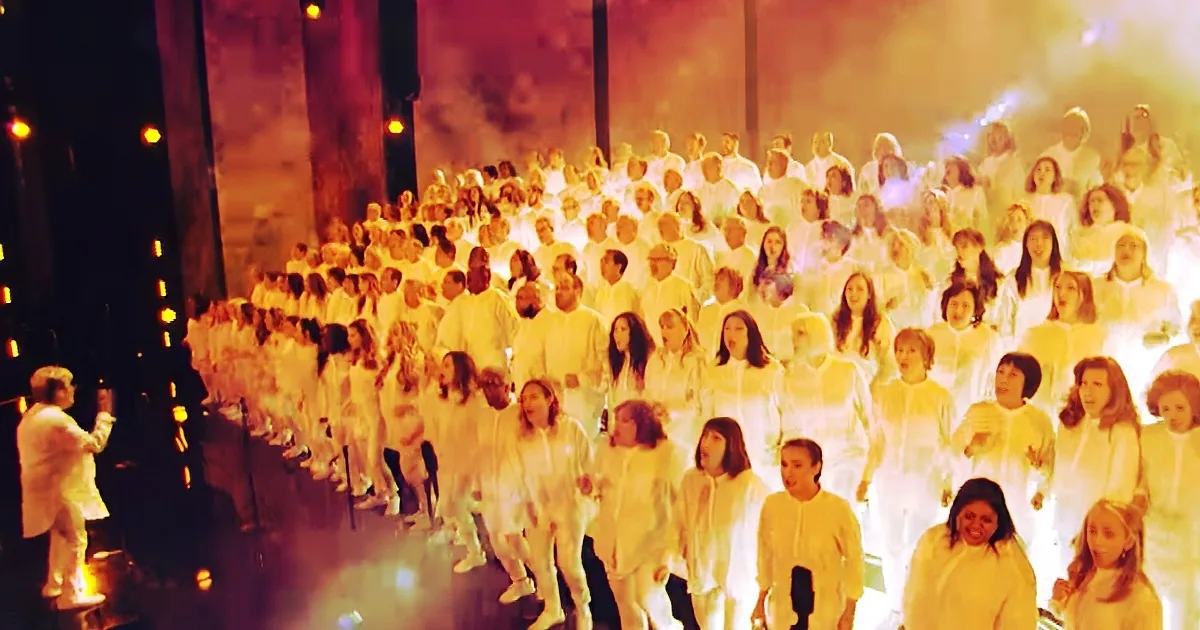 A choir in white robes sings under warm lighting, with a conductor leading from the left.