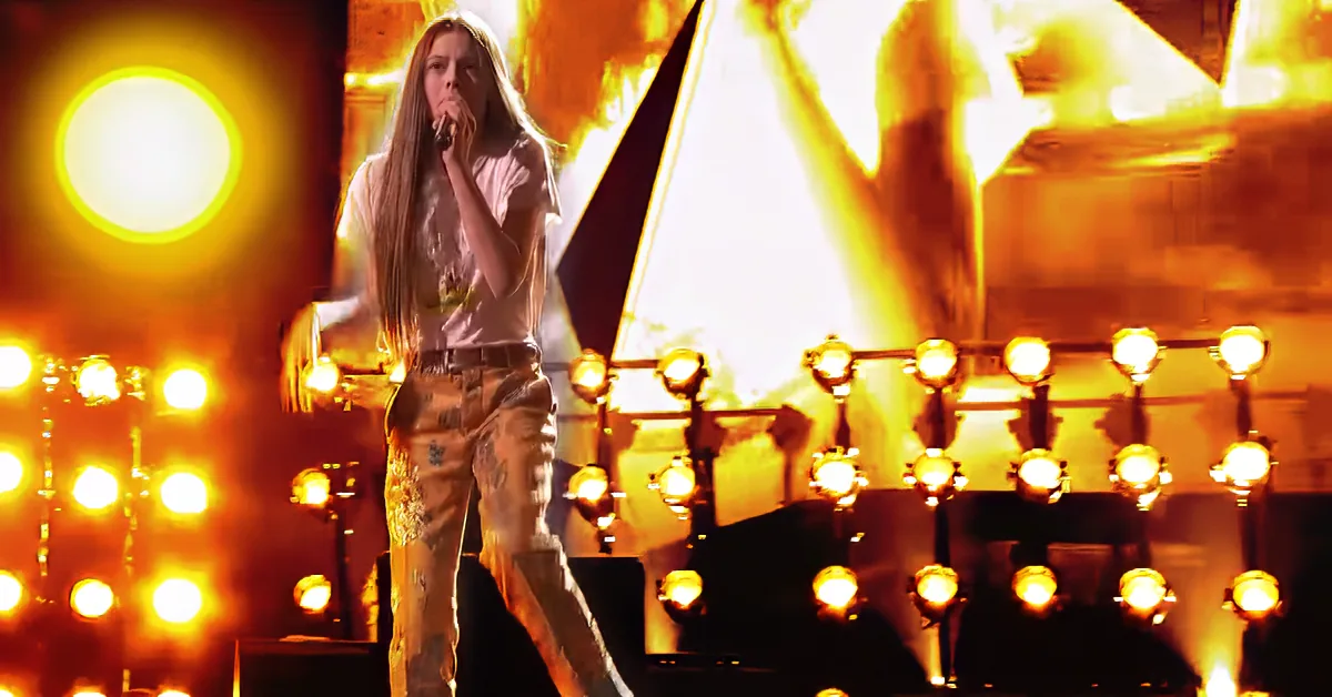 A person in a white shirt and patterned pants stands on stage with bright yellow lights and a large screen behind them.