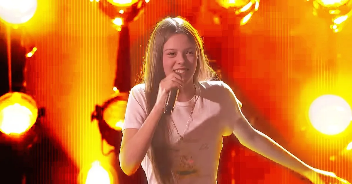 A person in a white shirt sings into a microphone on a stage with bright orange lights.