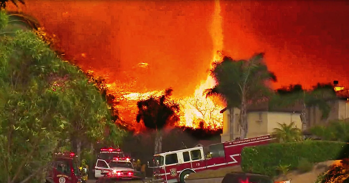 A massive wildfire engulfs a residential area, with intense flames and smoke, surrounded by emergency vehicles.