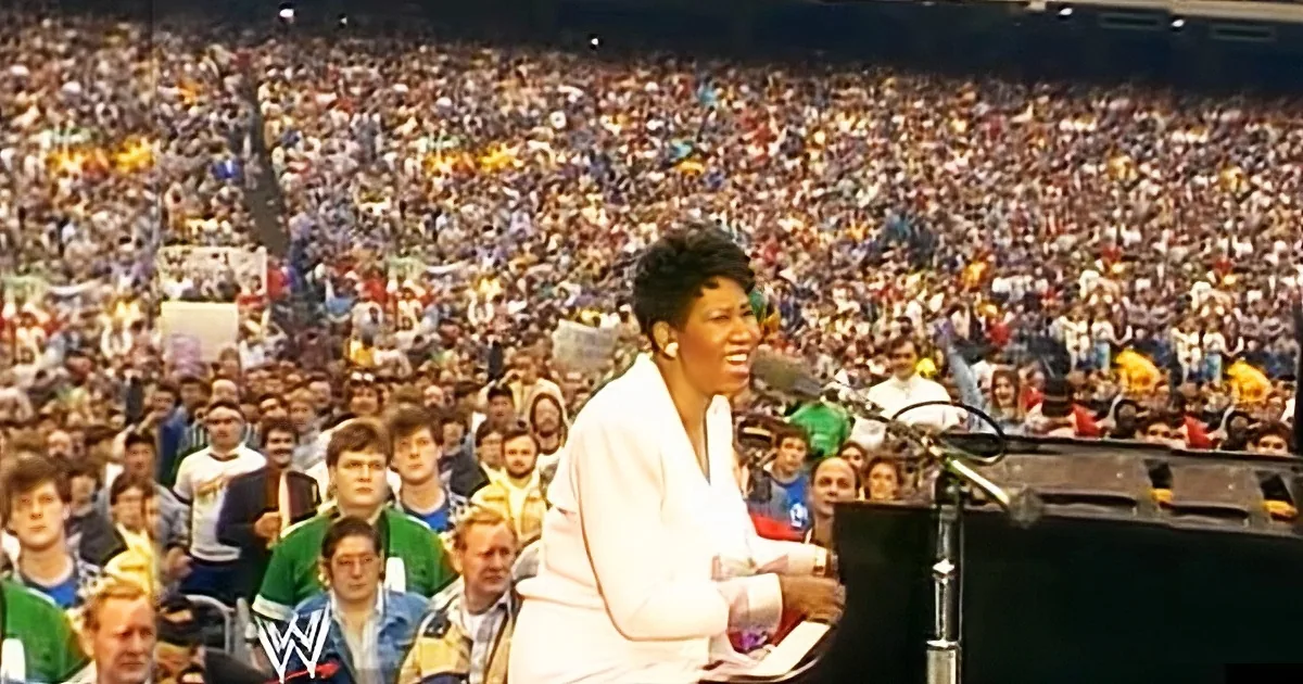 A woman in white stands at a podium, smiling, with a large crowd in the background.