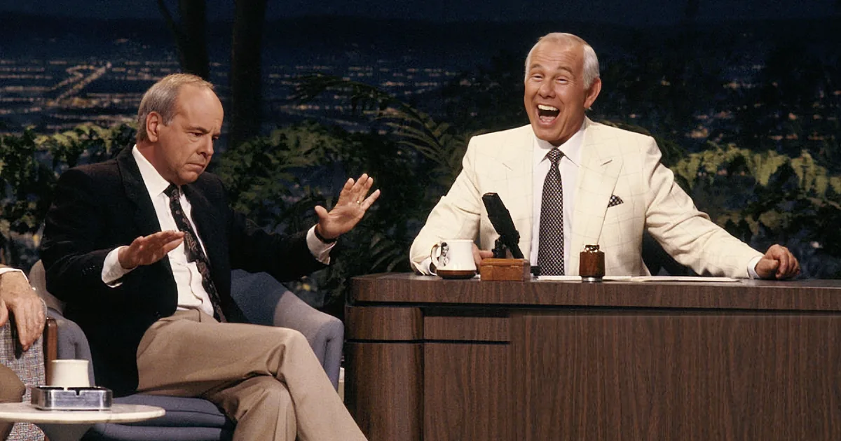 A man in a white suit sits at a desk, laughing, with another man in a suit gesturing beside him.