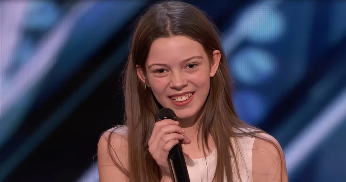A smiling young woman with long brown hair holds a microphone, wearing a light-colored top, against a blurred blue...
