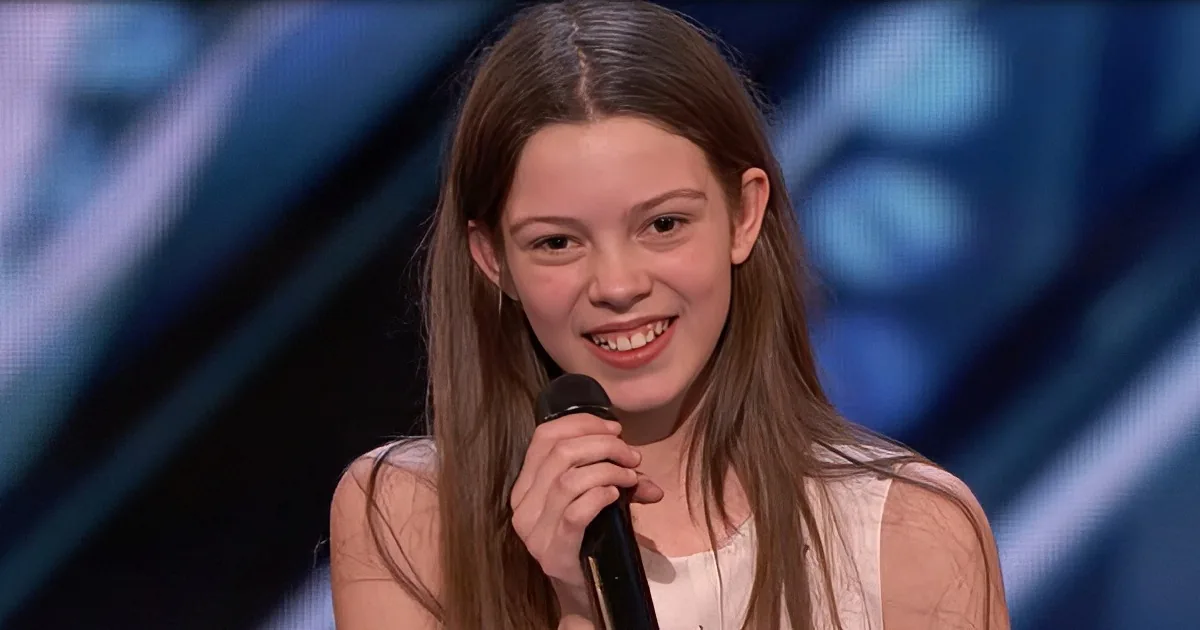 A young girl with long brown hair smiles while holding a microphone on stage.