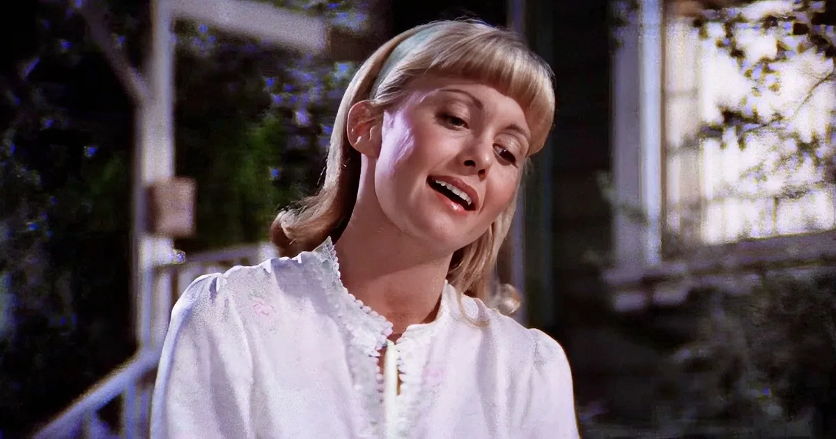 A woman with blonde hair in a white blouse stands outdoors, smiling with her mouth slightly open.