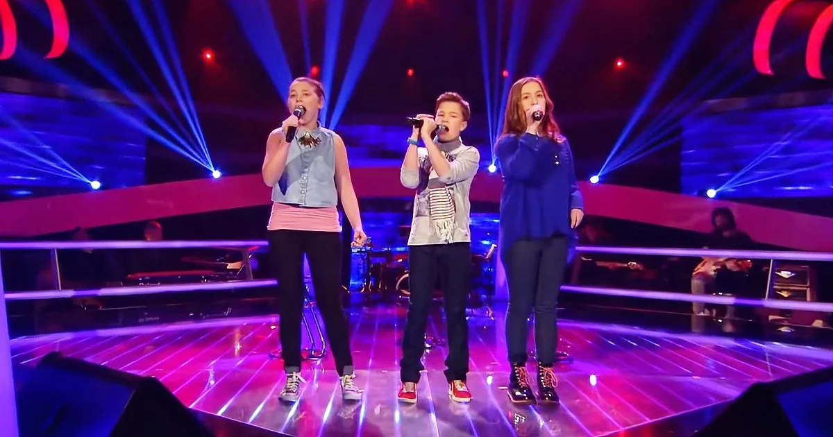 Three children sing on stage with microphones, illuminated by vibrant purple and blue lights.