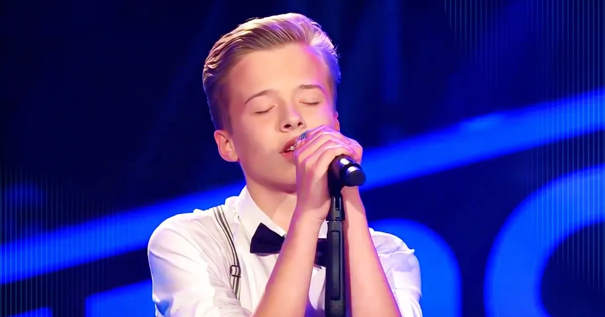 A young person in a white shirt and bow tie sings into a microphone on a stage with blue lighting.