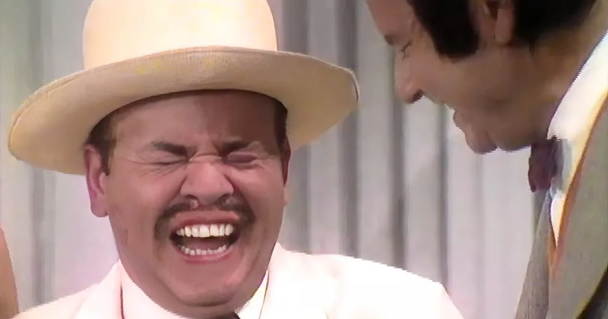 A man in a white suit and straw hat is laughing with another person nearby.