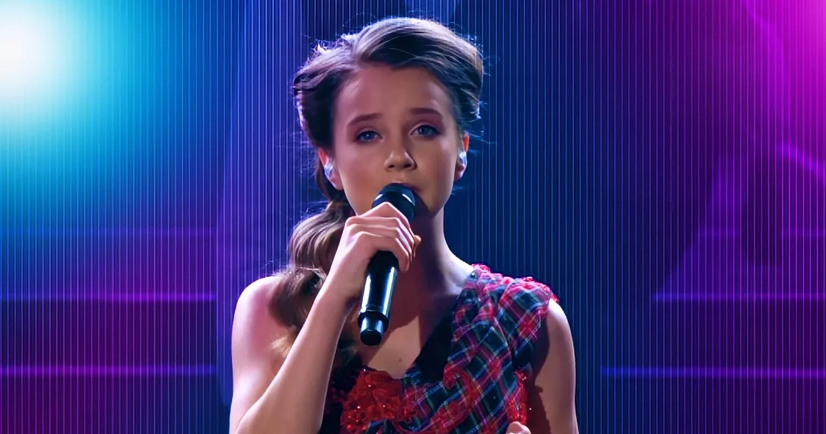 A person in a red plaid dress holds a microphone, singing against a vibrant blue and purple background.