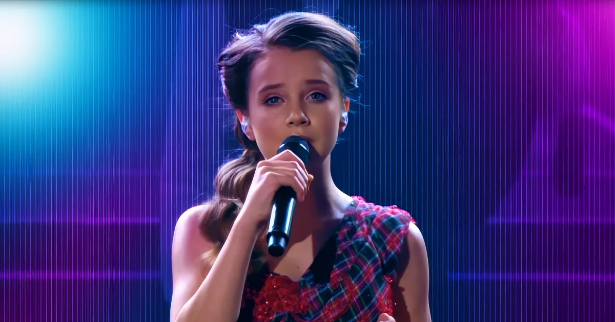 A young girl in a red plaid dress holds a microphone, singing on stage with colorful lights behind her.